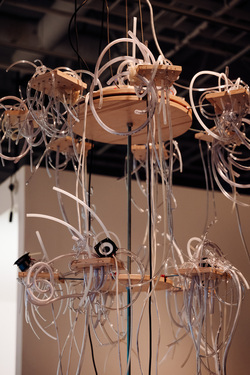 Multi-tiered suspended wooden chandelier with plastic tubes spilling outwards over the sides. There are four speakers and two microphones integrated into the sculpture.