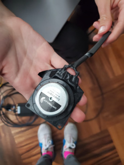 Close-up photo of a person's hands holding a black circular electronic device with a cable,with silver text against a black background which reads “DAYTONAUDIO” in uppercase with a logo of an arch consisting of 3 lines, and below that black text against a silver background reading “DAEX58FP,” “8 ohm/25W,” and “Made in China.” .