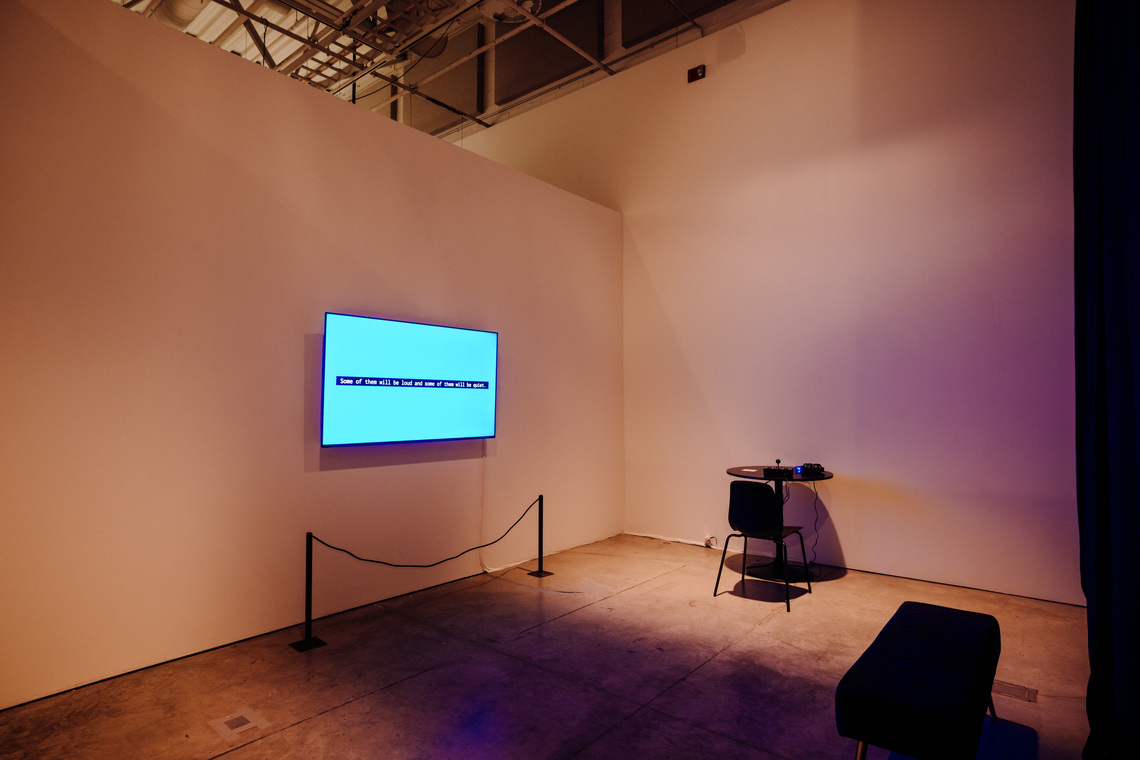 Room with a circular table against the back wall. A chair sits in front of the table, facing the wall. A large monitor displays text “Some of them will be loud and some of them will be quiet” Across from the monitor is a short bench.