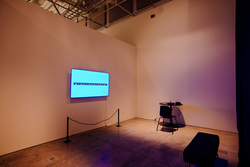 Room with a circular table against the back wall. A chair sits in front of the table, facing the wall. A large monitor displays text “Some of them will be loud and some of them will be quiet” Across from the monitor is a short bench.