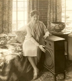 A fading photograph from the mid 1920s of a middle-aged woman wearing an elegant pleated skirt and draped sweater. She sits on a day-bed next to a wooden radio with curving legs, about three feet tall. One forearm rests on top of the radio; the other hand touches the front.