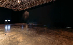 Interior of a dimly lit exhibition space with glossy concrete floors, dark painted walls, and an exposed ceiling structure. On the left, a set of screens display images, casting light on the floor.