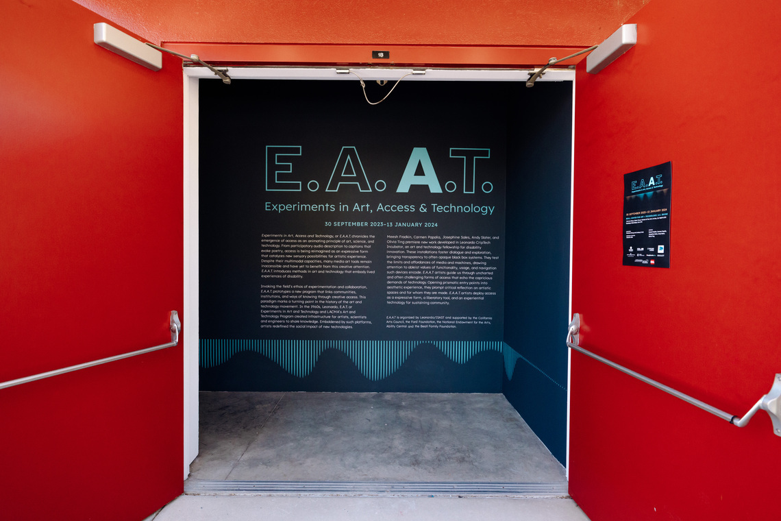 The exterior of the Beall Center for Art and Technology, where the exhibition is taking place. The building's bright red facade prominently displays raised metallic lettering reading "Don and Joan Beall Center for Art and Technology" along with the street number 712. Above, a large blue banner for the "E.A.A.T." exhibition is affixed to the wall. The entrance to an exhibition space has bold red double doors opened to reveal a dark blue wall inside. The exhibition is titled "E.A.A.T.: Experiments in Art, Access & Technology", running from September 30, 2023, to January 13, 2024. The large text on the wall introduces the exhibition. The wall features a modern, minimal design with blue-green typography and a waveform graphic at the bottom. A smaller informational sign is mounted on the right red door.