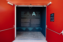 The exterior of the Beall Center for Art and Technology, where the exhibition is taking place. The building's bright red facade prominently displays raised metallic lettering reading "Don and Joan Beall Center for Art and Technology" along with the street number 712. Above, a large blue banner for the "E.A.A.T." exhibition is affixed to the wall. The entrance to an exhibition space has bold red double doors opened to reveal a dark blue wall inside. The exhibition is titled "E.A.A.T.: Experiments in Art, Access & Technology", running from September 30, 2023, to January 13, 2024. The large text on the wall introduces the exhibition. The wall features a modern, minimal design with blue-green typography and a waveform graphic at the bottom. A smaller informational sign is mounted on the right red door.