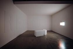 A gallery room with white walls, white ceiling, and a mottled concrete floor has a rectangular light source embedded in the right-side wall.  There are large raised capital letters R, E, S, C, U on the left-side and back walls, and a white rectangular block seating in the center of the room.