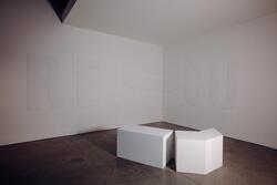 The corner of a gallery room with white walls and a mottled concrete floor. There are large raised capital letters R, E, S, C, U on the walls  and white rectangular block seating in the center of the room.