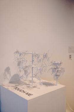 Two white sculptures atop a white rectangular pedestal in front of a white wall. Each sculpture consists of a dowel or pole with two regularly spaced circular platforms and a circular platform at the top. Thin white tubes cascade off the platforms in disarray. Black block letters on the pedestal say, “TOUCH ME.”