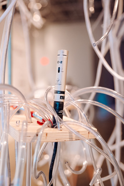 A Behringer C-2 condenser microphone is mounted on a wooden platform, surrounded by a dense network of clear plastic tubes. The microphone is securely fixed, with a black XLR cable running downward from its base. The wooden platform is supported by vertical rods and has various tubes weaving through and around it, some clamped with small red fasteners.

The background is softly blurred, with warm ambient lighting casting reflections on the tubes, creating a sense of depth.