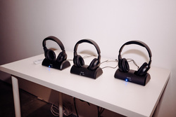 Three pairs of black wireless headphones ist on individual charging docks on a white table against a plain white wall. The headphone are available for visitors to experience Olivia Ting's Song without Words.

Each dock has a blue LED indicator light, suggesting that the headphones are powered on or charging. The headphones are identical in design, with padded ear cups and an adjustable headband.  Several black cables run along the back of the table, connecting the docks to a power source.