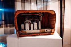 A vintage wooden radio casing is displayed on a white pedestal. The back panel of the radio is removed, exposing its internal electronic components, including metallic boxes, wiring, and a circular tuning mechanism. The wooden casing has a curved design with horizontal grooves along the interior, showing signs of age and wear.

There are small labels and markings on the internal components, including a serial code (2076112) and other technical specifications, suggesting that this is an authentic historical piece. Handwritten markings are visible on the bottom wooden edge of the radio.

The background consists of a softly lit exhibition space with translucent black curtains
