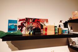 The image features a dark floating shelf mounted on a white wall, displaying a mix of books, self-care items, and personal essentials. On the left side, a green folded shirt rests beside a book titled Sea and Fog by Etel Adnan. Next to it, four comic books are standing upright: Daredevil: No Fear, Thor: Ragnarok, Astonishing X-Men, and The Walking Dead.To the right of the books, there is a fabric-covered tissue box with a red and green plaid pattern. Further right, personal care items are arranged, including a green glass cup with a straw, a bottle of eucalyptus massage oil, a container labeled Simple Blossom, a dropper bottle of glycogen supplement, and a bottle of Aveeno Stress Relief lotion.