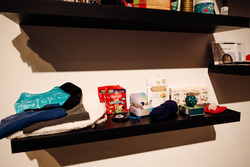 The image features a dark floating shelf mounted on a white wall, containing various self-care and comfort items. On the left side, there is a blue gel ice pack with black straps, stacked on top of folded soft fabric, including a white plush item and dark-colored clothing.In the center, there are two red bags of Maynards Wine Gums and a small blue tin with sewing needles and thread. Behind them, there's a pack of Dandelion Leaf & Root Tea. To the right, there are multiple boxes of Welly bandages in pastel-colored packaging.Further right, there is a soft navy blue sock, a green spiky massage ball, a small box of matches, and a red pouch with a translucent pink object on top. Above this shelf, another floating shelf is visible, holding personal care items, including massage oil and supplements.