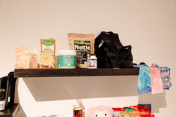 The image features a dark floating shelf mounted on a white wall, holding a variety of health and wellness products. Items on the shelf include Emergen-C vitamin C packets in tangerine flavor, a carton of Pacific Foods Organic Butternut Squash Soup, a container of Green Superfoods Collagen, a bottle of Magnesium Bisglycinate, and a brown paper bag labeled Nettle Leaves Cut & Sifted. To the right of these items, there is a black orthopedic or posture support brace. Next to it, there are two neatly folded fabric items with colorful patterns, one in blue swirls and the other in a pink and white design.