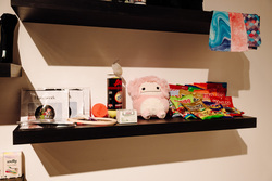 The image features a dark floating shelf mounted on a white wall, holding an assortment of personal and comfort items. On the left side of the shelf, there are handwork embroidery kits in clear plastic packaging, a pair of embroidery scissors, and a wooden embroidery hoop. Next to them, there's a small orange container labeled "Poem", a white box labeled "Cedar", and a tall canister of American Select Champion cue chalk with the lid open.In the center of the shelf sits a small pink plush toy resembling a fluffy creature with a neutral facial expression. To the right, there is a large collection of Haribo gummy candy packs, stacked in various colors and flavors.
