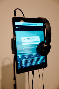 A tablet mounted on a stand, displaying a website called "Weather for the Blind." The screen features a blue-toned interface with a streaming audio player and descriptive text explaining the project, which appears to involve sonifying weather data. A pair of black Sony headphones is hooked over the top-right corner of the tablet, with their cable hanging down and plugged into the device. The stand holding the tablet has a metal arm extending behind it, and a headphone jack cable is visibly connected to the top left of the tablet. The background is a neutral-colored wall. The lighting is soft and even, highlighting the screen's content without harsh reflections.