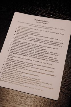 The image shows a printed document titled "Pain Pals Survey" by Carmen Papalia on a dark wooden surface. The page contains a series of reflective questions about pain, self-care, support systems, and societal perspectives on pain. The text is structured with bullet points, each prompting the reader to consider aspects such as how they describe pain, what brings comfort, and what a supportive environment might look like. The document has a slight curl at the top left corner. The lighting casts a soft shadow on the right side, emphasizing the texture of the paper and the dark wood grain underneath. The text is printed in a clean, readable font, and the document appears to be part of a take-home exercise encouraging deep personal reflection.
