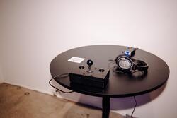 Circular table with joystick and buttons on a controller. There is a headphone amp with lights on and a pair of headphones directly in front of the controller. To the left of the controller is a QR code.