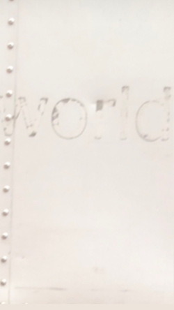 An image shows a pale, off-white piece of sheet metal with decal residue faintly spelling the word “world.” Screws line the metal’s left edge, emphasizing its industrial, institutional quality. This subtle, worn surface echoes themes in Rescue, where the plaster relief is designed to blend almost imperceptibly into its architectural surroundings, reflecting on visibility, erasure, and the muted presence of institutional structures.