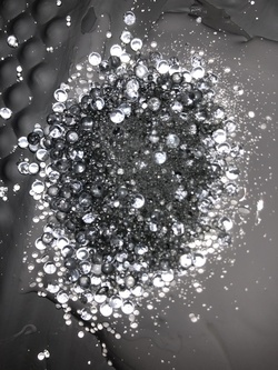 An image shows a smooth grey surface scattered with a pile of grey powder and transparent spherical beads of varying sizes. The beads appear to glow softly. This composition represents an early material exploration for Rescue, a shallow, floor-to-ceiling plaster relief that is part of the installation Total Running Time.
