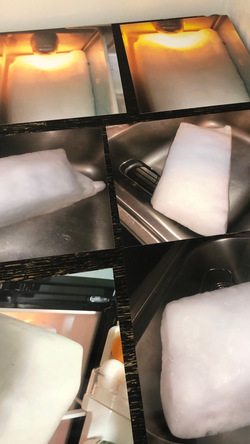 An image shows six photographs arranged on a piece of wood. Each photo features a large rectangular slab of ice placed in a sink or in front of a freezer. In the first two photographs, the ice slabs are bathed in a warm orange light, suggesting an early visual exploration for Day for Night, an artwork that employs two prepared 7,000 Kelvin circadian optic light panels commonly used to mimic natural sunlight.