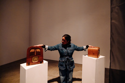 A dark-haired woman in a denim jumpsuit stands between two pedestals, gently resting her hands on two wooden vintage radios.