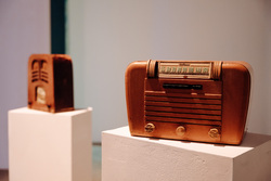 Two wooden vintage radios on white pedestals. The one in the background stands vertical, the one in the foreground is wide with three knobs.
