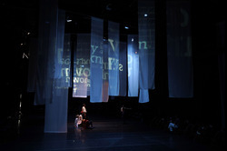 A dimly lit theater stage with translucent blue-tinted curtains hanging from above, some with yellow serif letters projected onto them. Performers are scattered on the stage, some standing and some seated on the floor, with one individual seated on a chair in the foreground to the right.