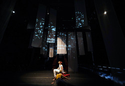 A line of five performers either kneeling or in the process of getting up to kneel on a dimly lift, amid vertical white banners displaying projected scattered text and closed captions reading “new student from Japan” in English and Japanese.