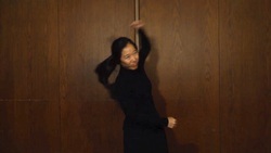 A person in a black outfit dancing with an expressive arm movement, raising their left hand,  against a plain wooden background.
