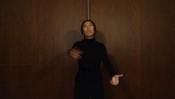 A person with arms in motion stands in front of a wooden wall, wearing a black long-sleeved top, looking upwards to the left with pursed lips.