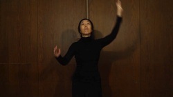 A person in black attire is standing against a wooden wall with an expressive pose, arms raised and one leg slightly lifted, suggesting a dance or performance in motion.
