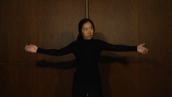 A person in a black turtleneck with arms spread in motion against a wooden wall looking to her left with a concerned expression.