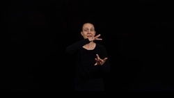 A person in black clothing appears to be using sign language against a dark background.