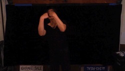 Person standing in front of a black background with their fists’ knuckles connected  over their face, a white sign labeled in black reading "GARAGE SALE" situated at the bottom left and a blue sign with white text reading “408 243-REAL” in a dimly lit setting.