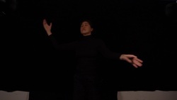 A person in black clothing with eyes closed and arms and hands outstretched in movement in a dark room.